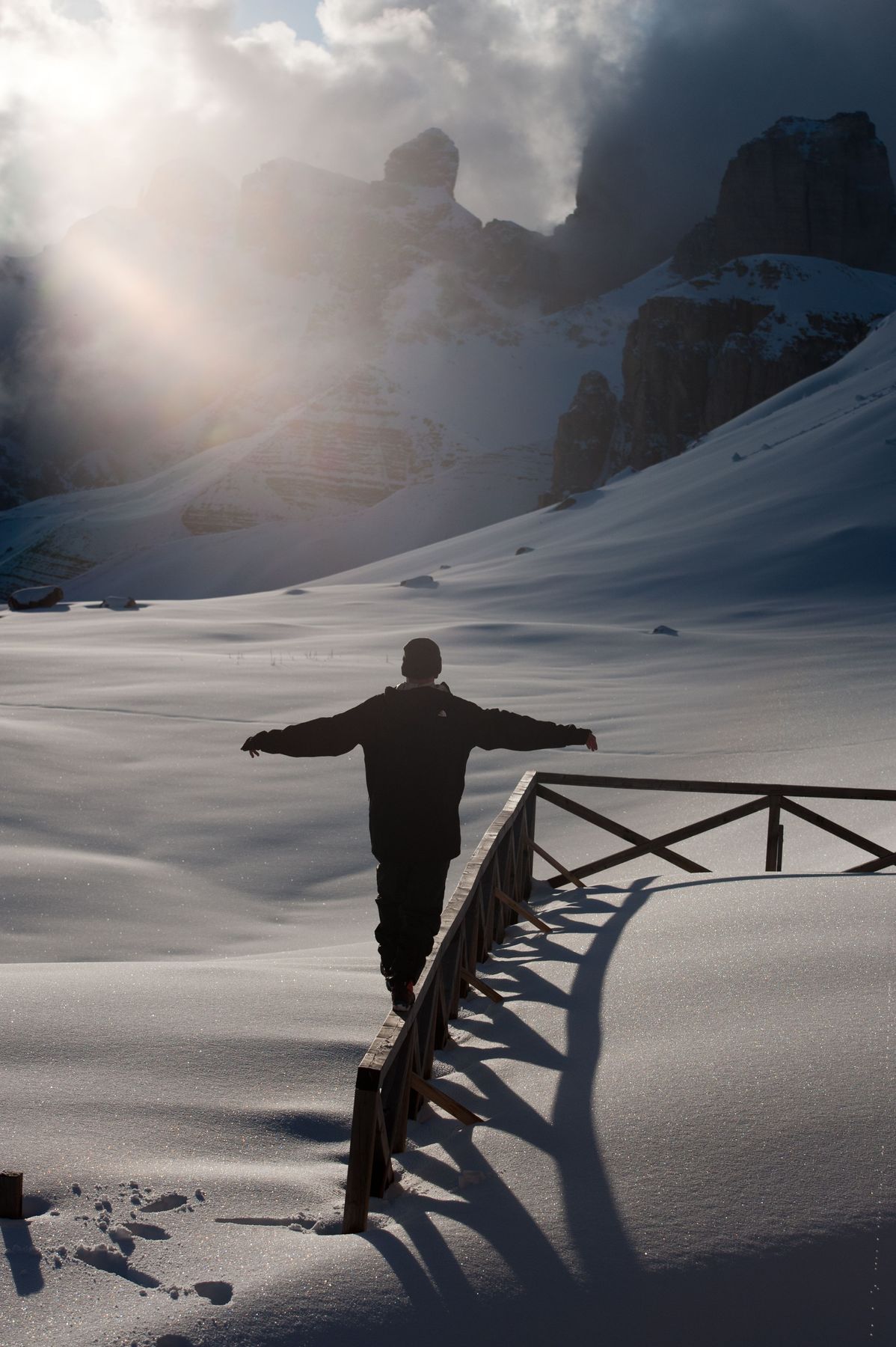 A person walks along a wooden fence in bright powder snow with sun breaking through cloud